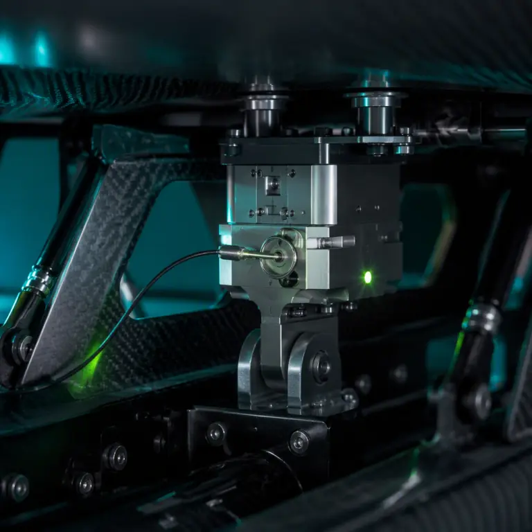 Professional photograph of a UAV payload bay on a dark carbon fiber airframe, close-up of a compact machined aluminum safe-and-arm interlock unit mounted between the airframe hardpoint and a payload release hook, with visible mechanical safety pin and a small status LED glowing lime green, teal and lime-green accent lighting, shallow depth of field, photorealistic, no people faces, no text, no logos