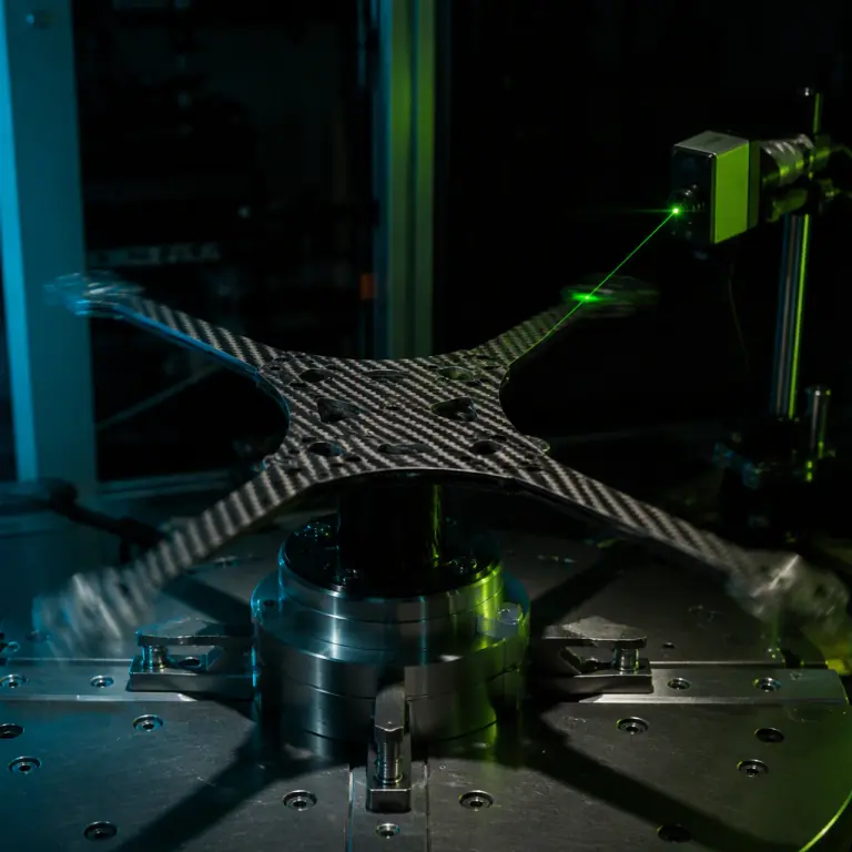 Photorealistic render of a carbon fiber UAV airframe under animated vibration testing, faint motion blur visible along the boom tips with the centre plate relatively static, laser displacement sensor aimed at an arm, dark laboratory setting with teal and lime green accent light, no people faces, no text, no logos
