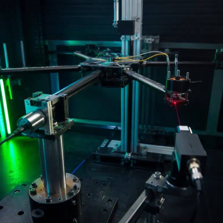 Photorealistic photograph of a UAV airframe mounted in a laboratory test fixture with a laser displacement sensor aimed at a motor mount and accelerometers cabled to the centre plate, load cell visible at the boom root, dark test cell with teal and lime green accent lighting, no people faces, no text, no logos
