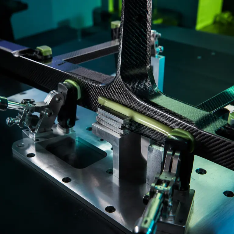 Photograph of a composite UAV frame assembly clamped in a machined bond jig on a dark bench, structural adhesive applied along a carbon fiber lap joint, rubber-tipped clamps and a shim pack visible at the interface, teal and lime green accent lighting, precision composites workshop aesthetic, photorealistic, no people faces, no text, no logos