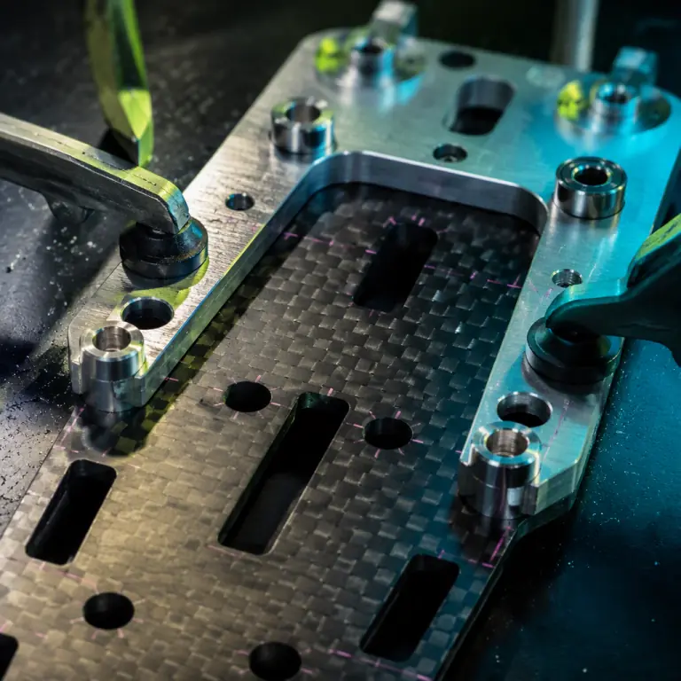 Close-up studio photograph of a machined aluminium drill jig plate with hardened steel bushings clamped to a glossy carbon fiber UAV centre plate on a dark workbench, precision layout blue dye marks visible on the composite, small swarf particles, lime green and teal rim lighting, photorealistic, no people faces, no text, no logos