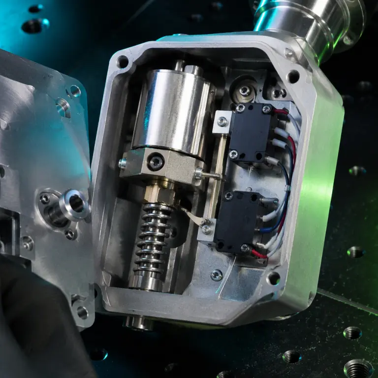 Macro studio photograph of a machined aluminum safe-and-arm housing partially opened on a dark anodized workbench, showing inside a rotary solenoid arm, a spring-loaded mechanical blocking pin, and two independent microswitches with fine wiring, precision aerospace hardware, teal and lime-green LED rim light, photorealistic, no people faces, no text, no logos