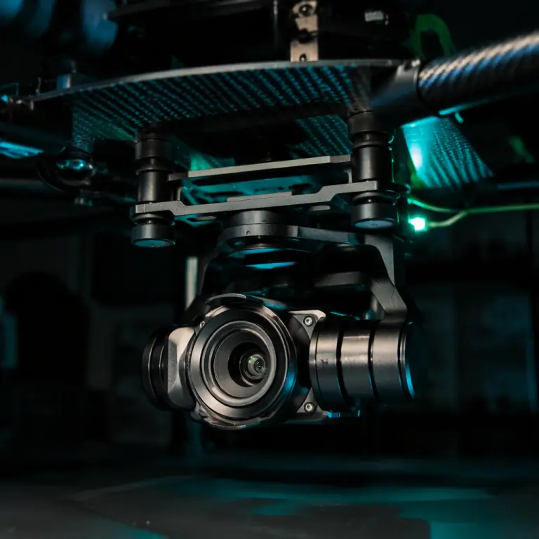 Photograph of a three-axis camera gimbal mounted under a carbon fiber UAV deck plate through four small cylindrical elastomer vibration isolators, dark engineering lab, precision-machined bracket, teal and green LED accent lighting, photorealistic, no people faces, no text, no logos
